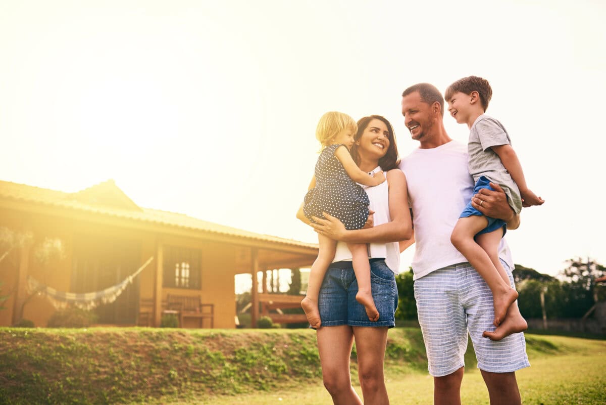 Smiling family with two young children standing in their sunny backyard in front of their new home, representing happy new homeowners in Richmond, Virginia.